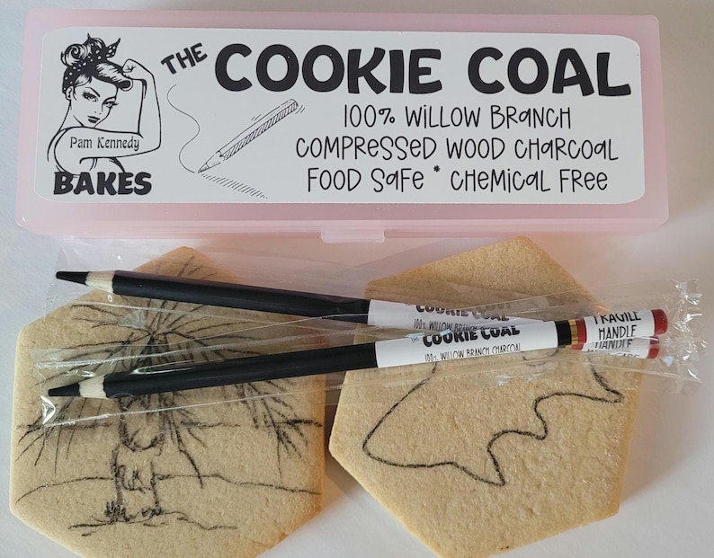 May include: A pink plastic box labeled "The Cookie Coal" with text and a drawing of a woman. Inside are black pencils and two cookies with drawings on them. The pencils are labeled "Cookie Coal".