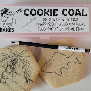 May include: A pink rectangular box with the text "THE COOKIE COAL" and "Pam Kennedy BAKES" sits above two decorated cookies and a black pencil. The pencil is labeled "COOKIE COAL" and "FRAGILE HANDLE". The cookies have drawings on them.