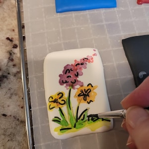 May include: A white, rectangular object with a floral design being painted. The flowers are yellow, purple, and pink with green stems and leaves. The object is on a metal rack.