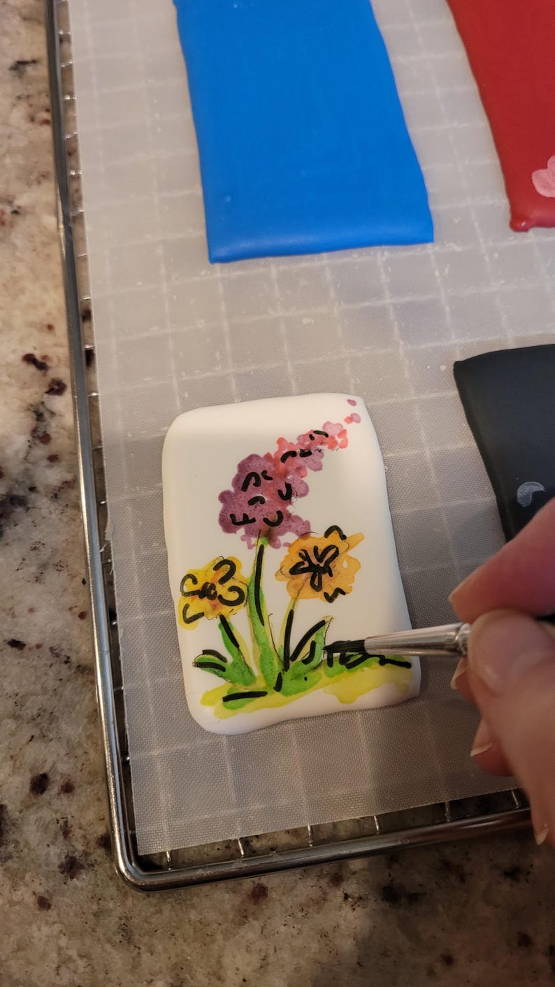 May include: A white, rectangular object with a floral design being painted. The flowers are yellow, purple, and pink with green stems and leaves. The object is on a metal rack.