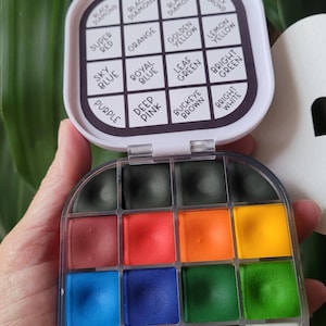 May include: A compact, open watercolor paint set with a variety of colors, including black, red, orange, yellow, blue, green, and purple. The lid displays color names like "Black Diamond" and "Super Red". The set is held in a hand.