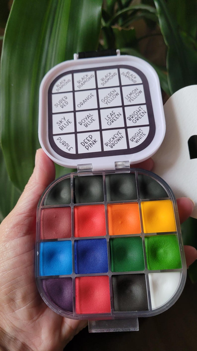 May include: A compact, open watercolor paint set with a variety of colors, including black, red, orange, yellow, blue, green, and purple. The lid displays color names like "Black Diamond" and "Super Red". The set is held in a hand.