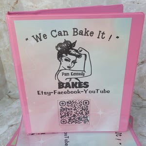 May include: Pink binders with the text "We Can Bake It!" and a logo for Pam Kennedy Bakes. The logo features a woman with her arm raised in a strong pose. The text "Etsy-Facebook-YouTube" is below the logo. A QR code is below the text.