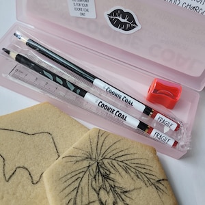 May include: A pink pencil case containing two pencils, a ruler, and a red pencil sharpener. The pencils and ruler are labeled "COOKIE COAL". Two square cookies with charcoal drawings are in the foreground. The case has the text "THE COOKIE COAL".