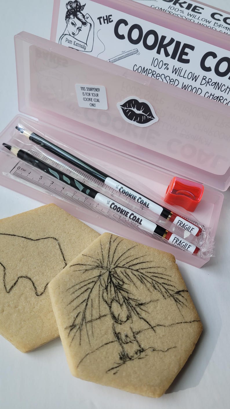 May include: A pink pencil case containing two pencils, a ruler, and a red pencil sharpener. The pencils and ruler are labeled "COOKIE COAL". Two square cookies with charcoal drawings are in the foreground. The case has the text "THE COOKIE COAL".