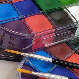 May include: A multi-colored face paint palette with a variety of colors including red, orange, green, and blue. Two paint brushes with silver handles and yellow bristles are in the foreground. The palette is in a clear plastic container.