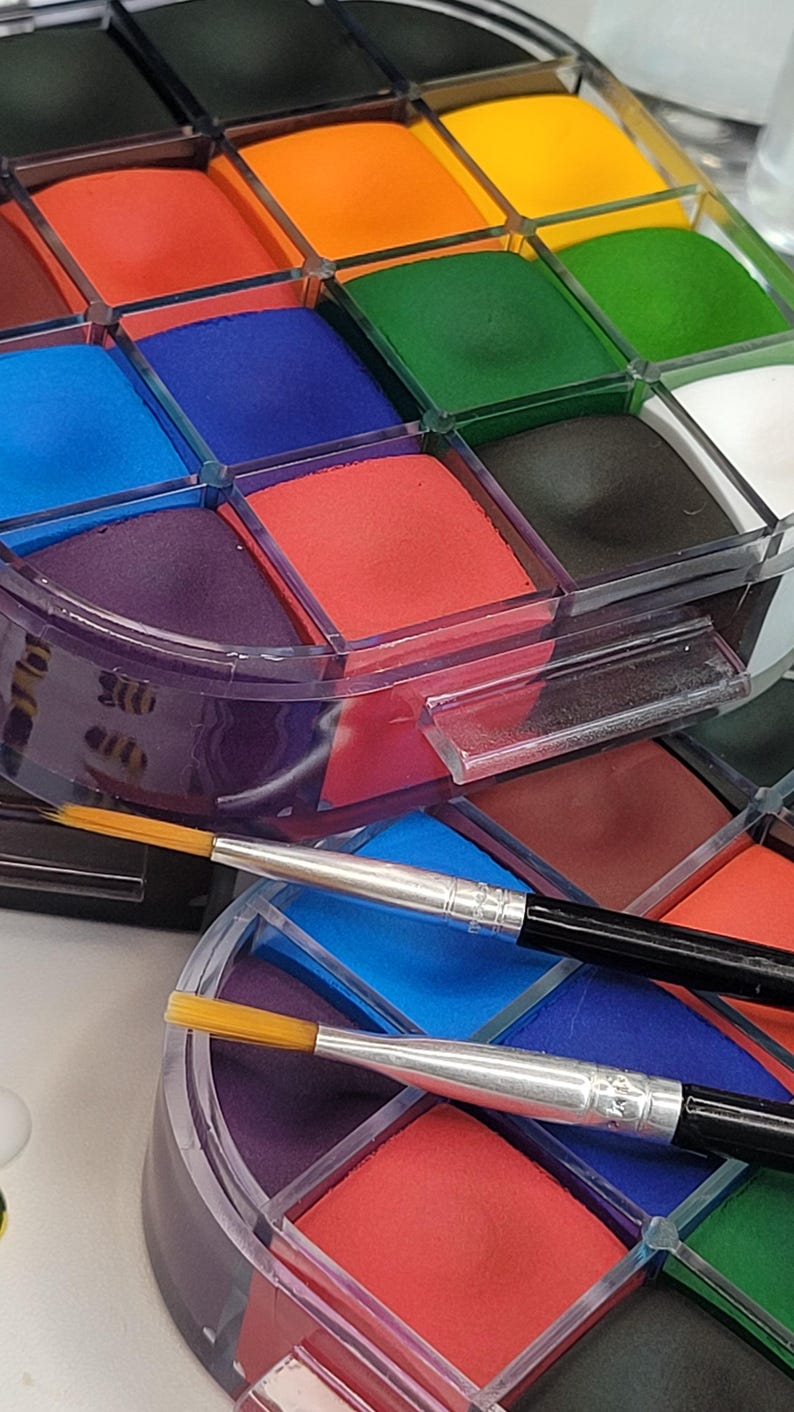 May include: A multi-colored face paint palette with a variety of colors including red, orange, green, and blue. Two paint brushes with silver handles and yellow bristles are in the foreground. The palette is in a clear plastic container.