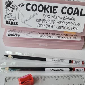 May include: Pink plastic boxes labeled "The Cookie Coal" with text, "100% Willow Branch Compressed Wood Charcoal Food Safe Chemical Free." Two pencils and a red pencil sharpener are visible. A ruler is also present.