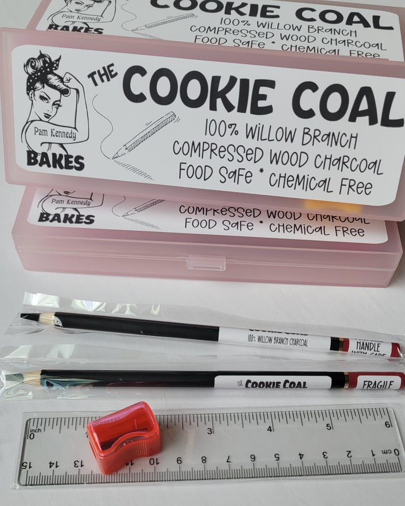 May include: Pink plastic boxes labeled "The Cookie Coal" with text, "100% Willow Branch Compressed Wood Charcoal Food Safe Chemical Free." Two pencils and a red pencil sharpener are visible. A ruler is also present.