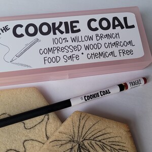 Peut inclure: Une bo&icirc;te rose rectangulaire avec le texte "THE COOKIE COAL" et "Pam Kennedy BAKES" est pr&eacute;sent&eacute;e. Un crayon noir et blanc marqu&eacute; "COOKIE COAL" et "FRAGILE" est pos&eacute; sur deux biscuits d&eacute;cor&eacute;s. Le crayon sert &agrave; dessiner sur les biscuits.