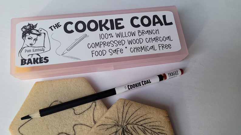 Peut inclure: Une bo&icirc;te rose rectangulaire avec le texte "THE COOKIE COAL" et "Pam Kennedy BAKES" est pr&eacute;sent&eacute;e. Un crayon noir et blanc marqu&eacute; "COOKIE COAL" et "FRAGILE" est pos&eacute; sur deux biscuits d&eacute;cor&eacute;s. Le crayon sert &agrave; dessiner sur les biscuits.