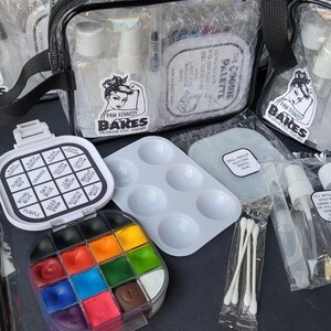 May include: A collection of art supplies, including paint palettes, brushes, and clear bags with spray bottles. The image features a variety of colorful paints and a reusable practice sheet. The bags have a black strap and a logo with a woman's face.