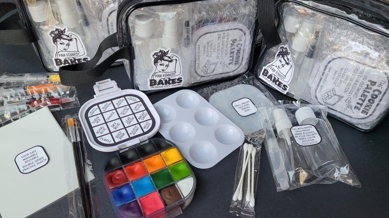 May include: A collection of art supplies, including paint palettes, brushes, and clear bags with spray bottles. The image features a variety of colorful paints and a reusable practice sheet. The bags have a black strap and a logo with a woman's face.