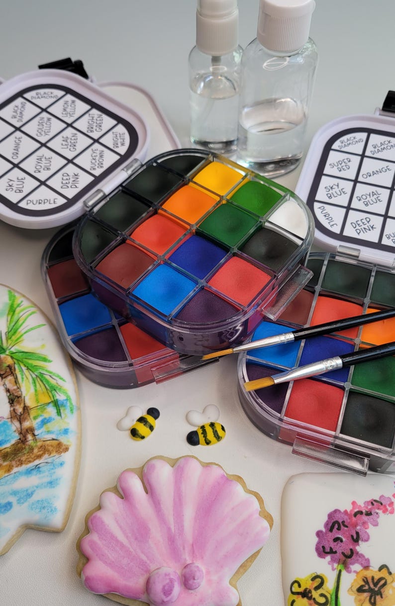 May include: An assortment of colorful face paint palettes with brushes, small spray bottles, and decorated cookies. The palettes contain various shades of colors, including orange, blue, and green. The cookies are decorated with a palm tree, bees, and a seashell.