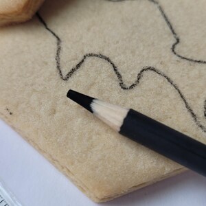 May include: Close-up of a black pencil with a white tip resting on a light-colored, rectangular cookie. A black outline of a design is drawn on the cookie. A ruler is visible in the lower left corner.