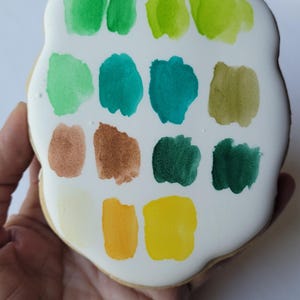 May include: A hand holds a cookie decorated with a white glaze and colorful paint swatches. The swatches include shades of green, teal, brown, yellow, and a pale yellow. The cookie has a rounded, irregular shape.