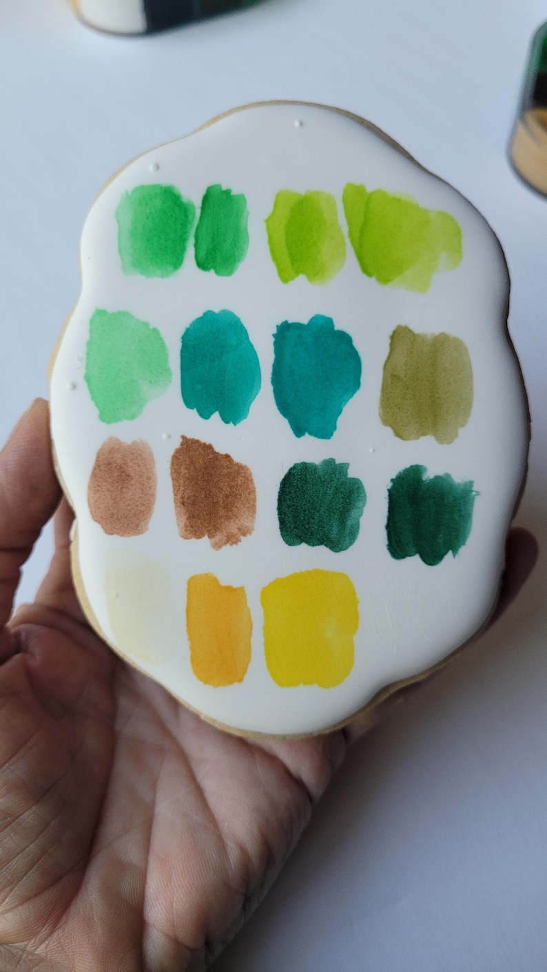 May include: A hand holds a cookie decorated with a white glaze and colorful paint swatches. The swatches include shades of green, teal, brown, yellow, and a pale yellow. The cookie has a rounded, irregular shape.