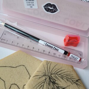 Peut inclure: Une trousse rose contenant une r&egrave;gle, un crayon Cookie Coal noir et blanc, un taille-crayon rouge et deux biscuits avec des dessins au fusain. La trousse porte l'inscription "The Cookie Coal".