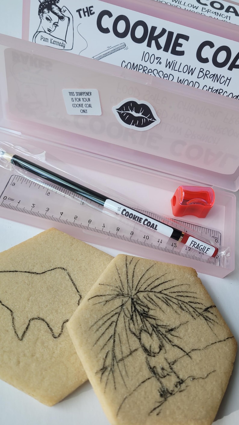 Peut inclure: Une trousse rose contenant une r&egrave;gle, un crayon Cookie Coal noir et blanc, un taille-crayon rouge et deux biscuits avec des dessins au fusain. La trousse porte l'inscription "The Cookie Coal".