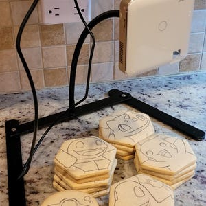 May include: A black adjustable stand holds a white and gold device. Several sugar cookies are decorated with cartoon character outlines. A black pencil rests on the counter. A power strip is mounted on the wall.