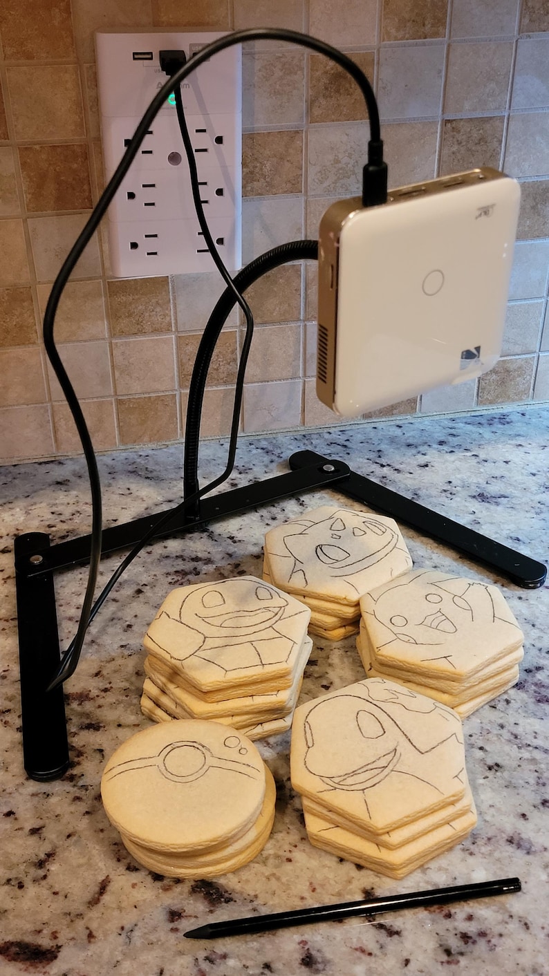 May include: A black adjustable stand holds a white and gold device. Several sugar cookies are decorated with cartoon character outlines. A black pencil rests on the counter. A power strip is mounted on the wall.