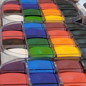 May include: A close-up view of several rows of colorful ink pads. The ink pads are arranged in a rainbow of colors, including red, orange, yellow, green, blue, and purple. The ink pads are in clear plastic containers.
