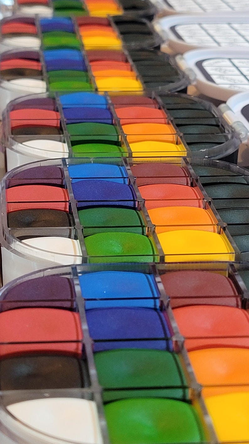 May include: A close-up view of several rows of colorful ink pads. The ink pads are arranged in a rainbow of colors, including red, orange, yellow, green, blue, and purple. The ink pads are in clear plastic containers.