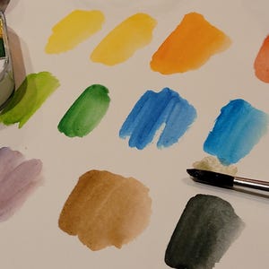 May include: A watercolor paint set with a variety of colors, including yellow, orange, blue, and green. Swatches of each color are painted on white paper. A paintbrush is also present, ready for use.