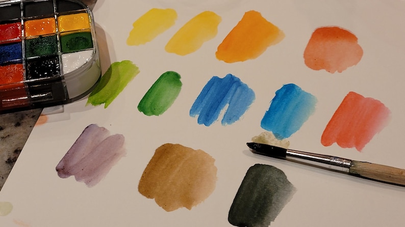 May include: A watercolor paint set with a variety of colors, including yellow, orange, blue, and green. Swatches of each color are painted on white paper. A paintbrush is also present, ready for use.
