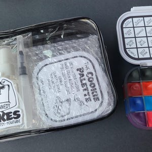 May include: A clear, zippered pouch containing a spray bottle, a syringe, and a sticker with the text "Pam Kennedy Bakes." Next to it is a watercolor palette with 12 colors and a lid with color names.