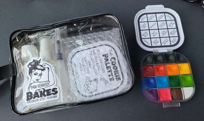 May include: A clear, zippered pouch containing a spray bottle, a syringe, and a sticker with the text "Pam Kennedy Bakes." Next to it is a watercolor palette with 12 colors and a lid with color names.