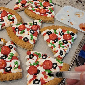 May include: Several pizza-shaped cookies with red tomato, black olive, and green pepper toppings. A watercolor paint set, palette, and a water brush are also visible. The cookies are on a baking sheet.