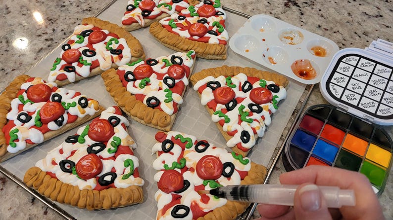 May include: Several pizza-shaped cookies with red tomato, black olive, and green pepper toppings. A watercolor paint set, palette, and a water brush are also visible. The cookies are on a baking sheet.