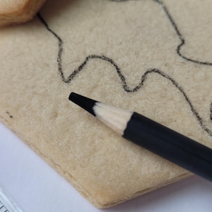 Peut inclure: Gros plan d'un crayon noir &agrave; pointe blanche pos&eacute; sur un biscuit rectangulaire clair. Un contour noir d'un dessin est trac&eacute; sur le biscuit. Une r&egrave;gle est visible dans le coin inf&eacute;rieur gauche.