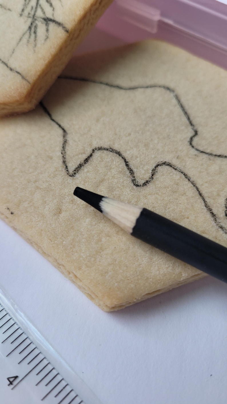 Peut inclure: Gros plan d'un crayon noir &agrave; pointe blanche pos&eacute; sur un biscuit rectangulaire clair. Un contour noir d'un dessin est trac&eacute; sur le biscuit. Une r&egrave;gle est visible dans le coin inf&eacute;rieur gauche.