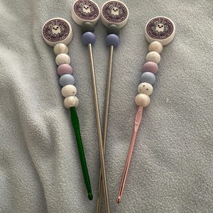 Puede incluir: Cuatro ganchillos con adornos decorativos. Los adornos presentan un diseño circular con una ilustración de fantasma y el texto "Living the Dream Nightmare at a Time". Los ganchillos tienen una variedad de colores, incluyendo verde, plateado y rosa.