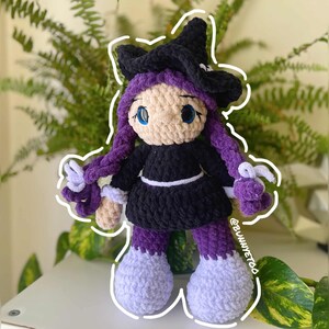 May include: A handmade crochet doll in the shape of a witch. The doll has a black witch hat, black dress, purple hair and leggings, and light purple shoes. The doll has blue eyes and a light skin tone. The doll is sitting on a white surface.
