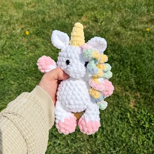 May include: A handmade white crochet unicorn with a yellow horn, pink hooves, and a colourful mane. The plush toy is soft and cuddly, perfect for children or as a decorative item. The unicorn is shown against a green grassy background.
