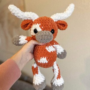 May include: A plush, orange and white crocheted longhorn steer. The stuffed animal has large white horns, white patches, and gray hooves and snout. The steer is soft and cuddly, perfect for a child's toy or nursery decor.