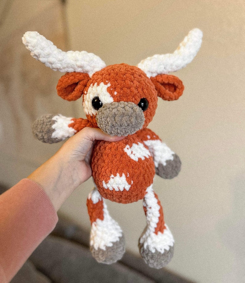 May include: A plush, orange and white crocheted longhorn steer. The stuffed animal has large white horns, white patches, and gray hooves and snout. The steer is soft and cuddly, perfect for a child's toy or nursery decor.