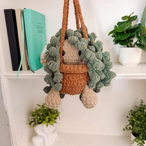 Op de afbeelding: Een handgemaakte, pluche hangende plantenbak met een grillig ontwerp. De plantenbak heeft een bruine pot met een lichtbruin gezicht en voeten, en groene, pluizige wol die op bladeren lijkt. De plantenbak wordt opgehangen door twee bruine koorden.