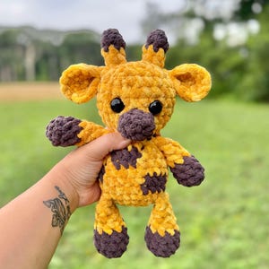 May include: A handmade crochet giraffe plush toy. The giraffe is primarily yellow with brown accents on the nose, hooves, and spots. It has black safety eyes and small ears. The plush is held up against a blurred green background.