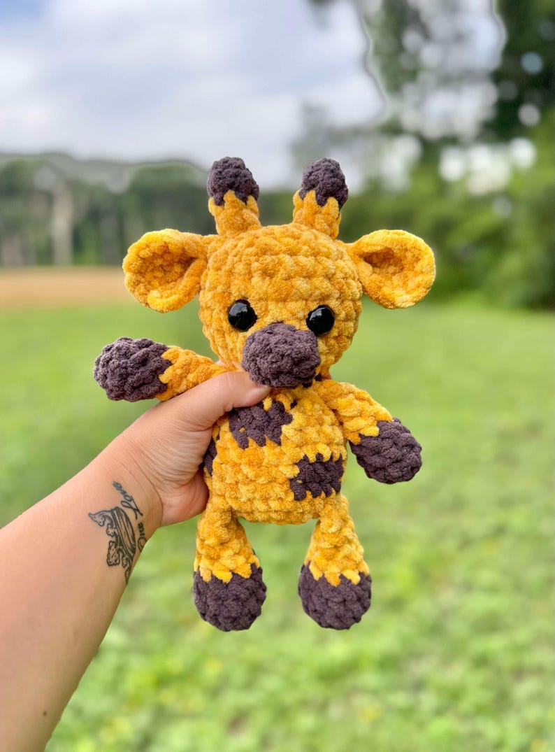 May include: A handmade crochet giraffe plush toy. The giraffe is primarily yellow with brown accents on the nose, hooves, and spots. It has black safety eyes and small ears. The plush is held up against a blurred green background.