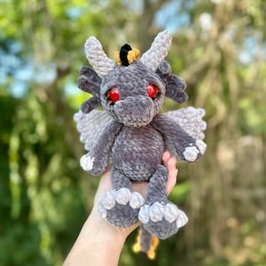 May include: A gray crocheted dragon with red eyes and a yellow crown. The dragon has wings and is sitting on a hand.