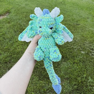 May include: A handmade crochet dragon plushie in shades of teal, blue, and white. The dragon has detailed wings, a long tail, and small, dark eyes. The plushie is held against a backdrop of green grass.