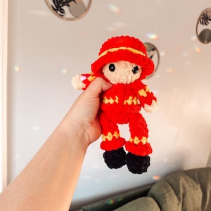 May include: A handmade crochet doll in a firefighter's outfit. The doll is mainly red with yellow details and black boots. It wears a red hat with a yellow trim. The doll is held up by a person.