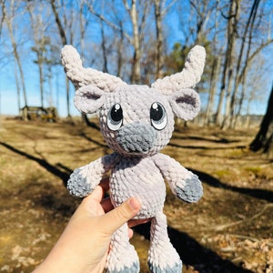 May include: A handmade, plush, light gray and gray stuffed animal with large, cartoon-style eyes and curved horns. The soft toy has a gray muzzle and feet. The background is a blurred outdoor scene with trees and sunlight.