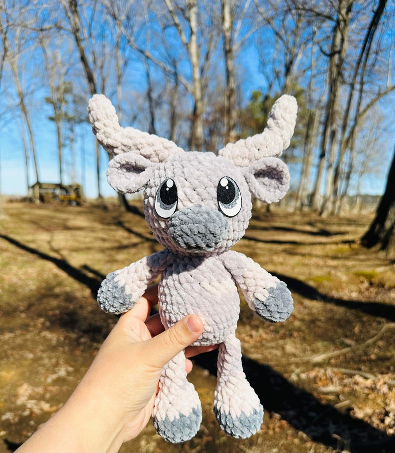 May include: A handmade, plush, light gray and gray stuffed animal with large, cartoon-style eyes and curved horns. The soft toy has a gray muzzle and feet. The background is a blurred outdoor scene with trees and sunlight.