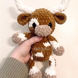 May include: A brown and white crocheted plush toy cow with white horns. The cow has a brown nose and black eyes. The toy is being held in a person's hand. @madelinemaeco