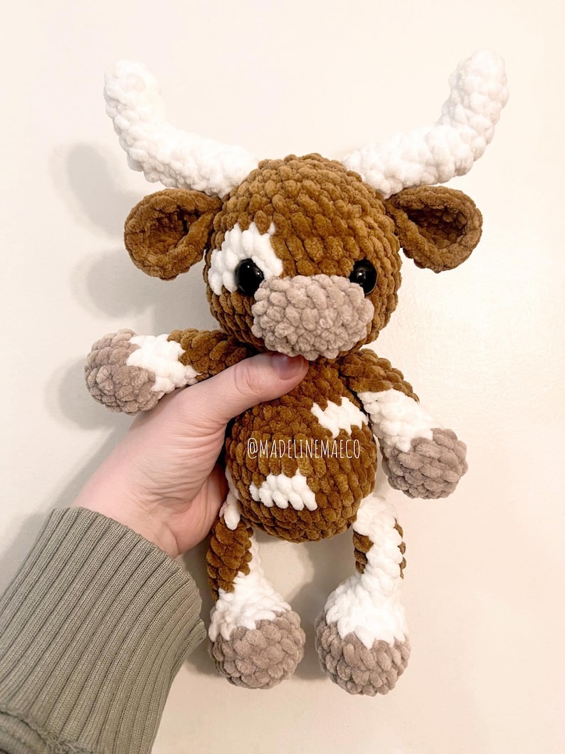 May include: A brown and white crocheted plush toy cow with white horns. The cow has a brown nose and black eyes. The toy is being held in a person's hand. @madelinemaeco